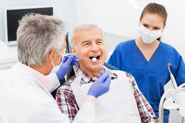 Picture of an older man with white hair, sitting in the dental chair and the dentist is completing an exam while the dental hygienist looks over his shoulder.