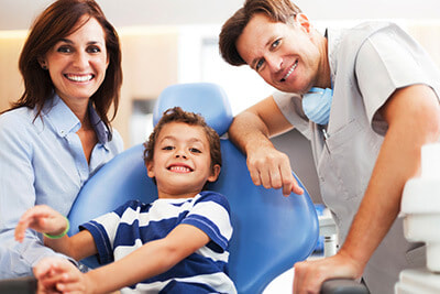 Photo of a Mother and her young son who is sitting in the dental chair.  The dentist is leaning over the patient and smiling.